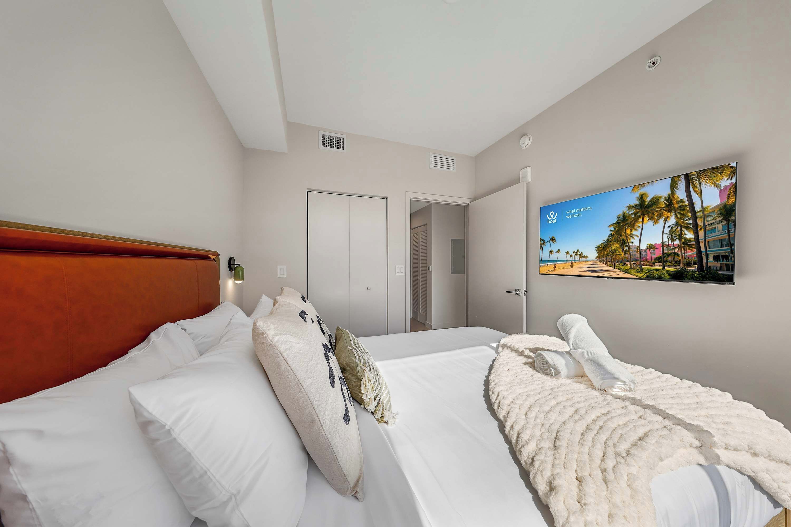 A hotel room with a bed, white bedding, a headboard, a wall-mounted TV, and a door leading to another room.