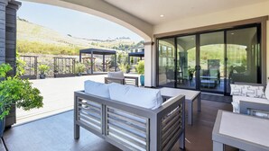 A covered outdoor patio with seating, a glass door leading to a garden, and a view of hills.