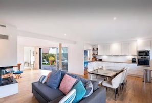 A modern living room with a sofa, a dining table, and chairs. There is a kitchen with white cabinets and appliances.
