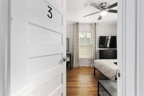 Ein Schlafzimmer mit Bett, Fernseher, Deckenventilator und einem Fenster mit Vorhängen.