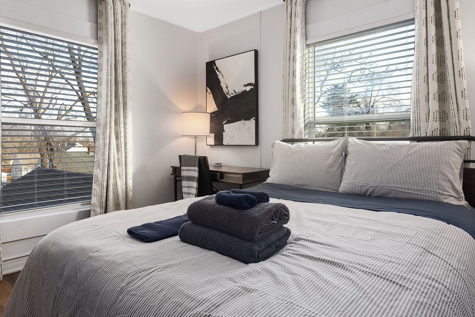 Une chambre d’hôtel équipée d’un lit, d’un bureau, d’une chaise, d’une lampe et d’une grande fenêtre avec des stores.