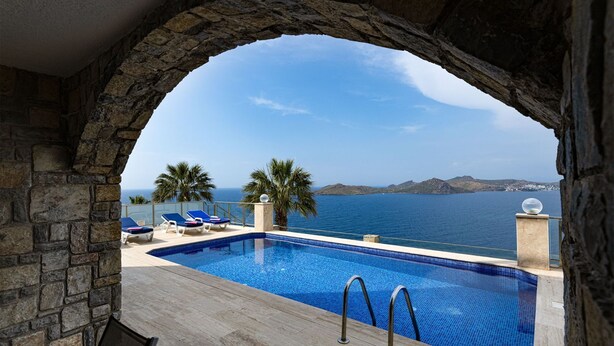 A pool with clear blue water, surrounded by stone walls and palm trees, overlooking a calm sea and distant hills. - BD4148 (Bodrum)