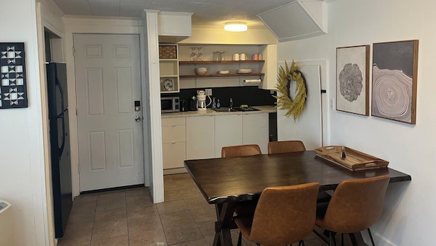 A dining area with a dark wooden table and brown chairs, a kitchen with white cabinets and a black countertop, and two framed artworks on the wall.