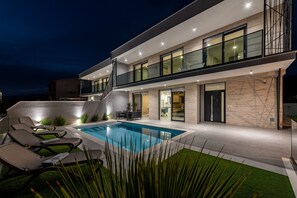 Een modern huis met twee verdiepingen, een zwembad, buitenmeubilair en een trap naar de bovenverdieping.