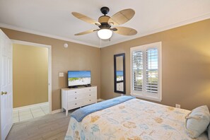Une chambre à coucher comprenant un lit, une commode, un téléviseur, un ventilateur de plafond, une fenêtre avec des stores et une porte.