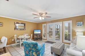 A living room with a television, a ceiling fan, a dining area with chairs, and a view through the glass doors. - Condo Near Beach with Pool PKRM (Pensacola)