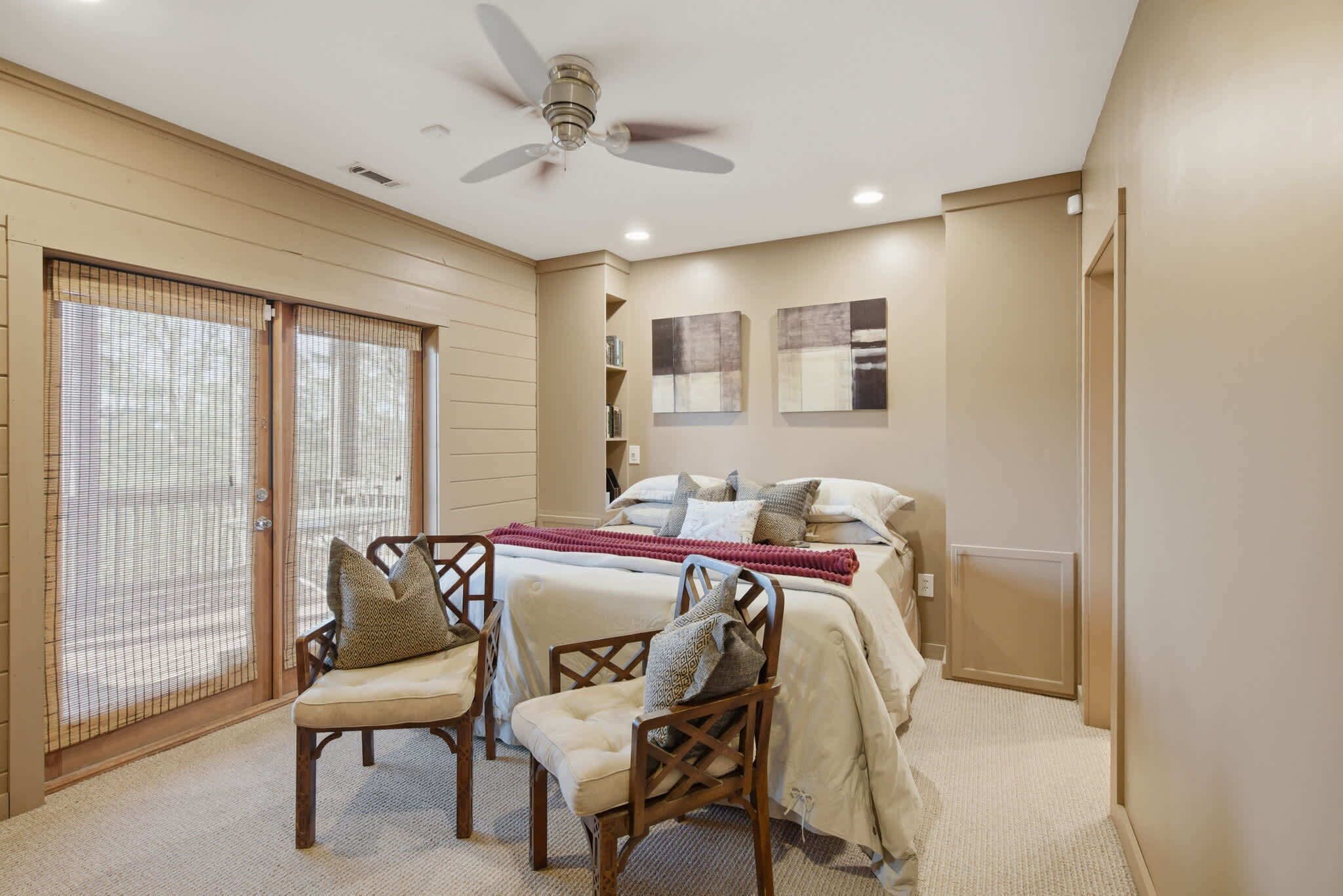A bedroom with a bed, two chairs, a ceiling fan, and a sliding glass door.