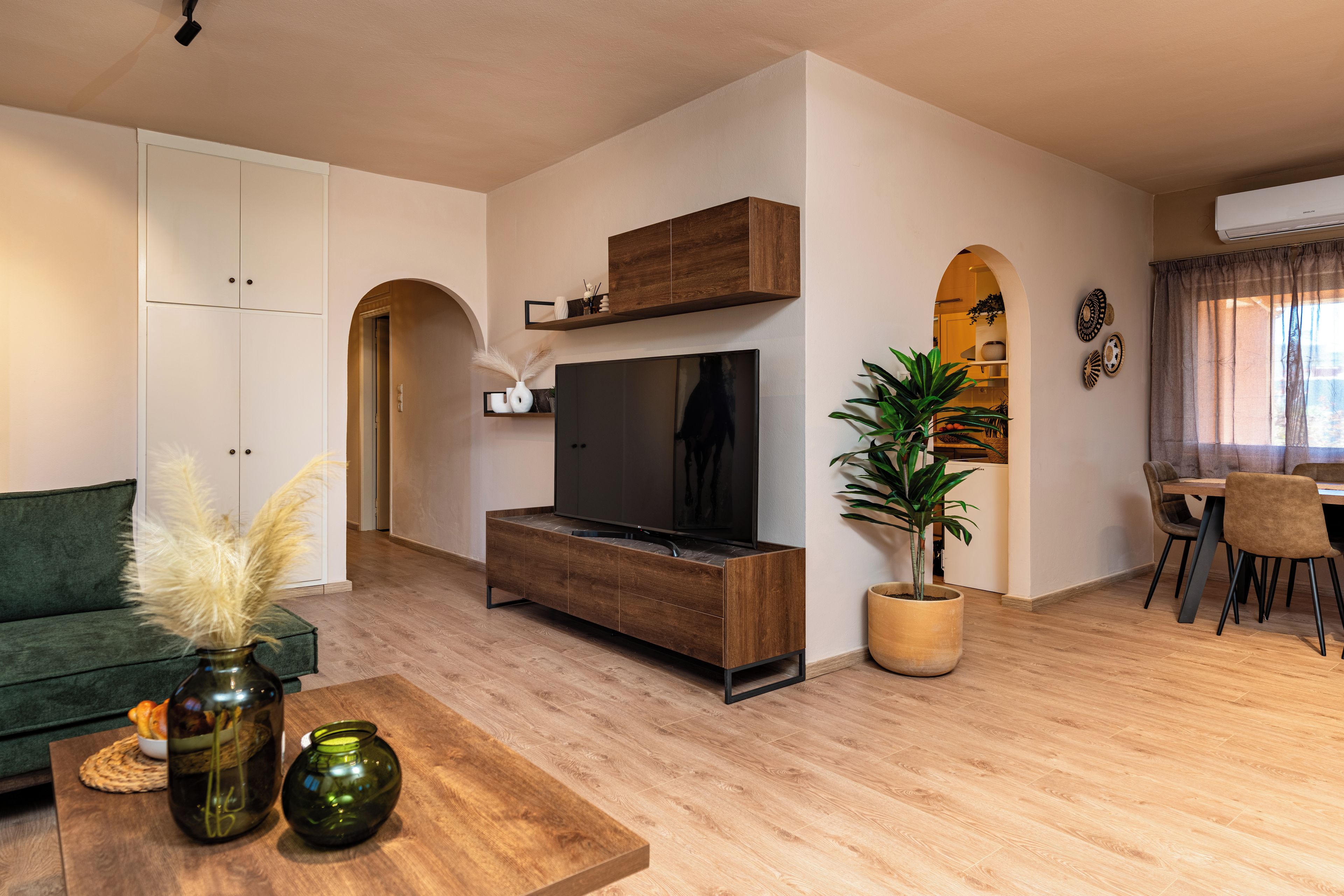 A modern living room with a flat-screen TV, wooden cabinet, green sofa, and a dining area with a table and chairs.