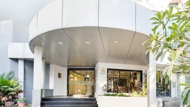 A modern building entrance with a curved overhang, steps leading up to a glass door, and potted plants around the area. - Super Deluxe Room Palette Country Club (Surat)