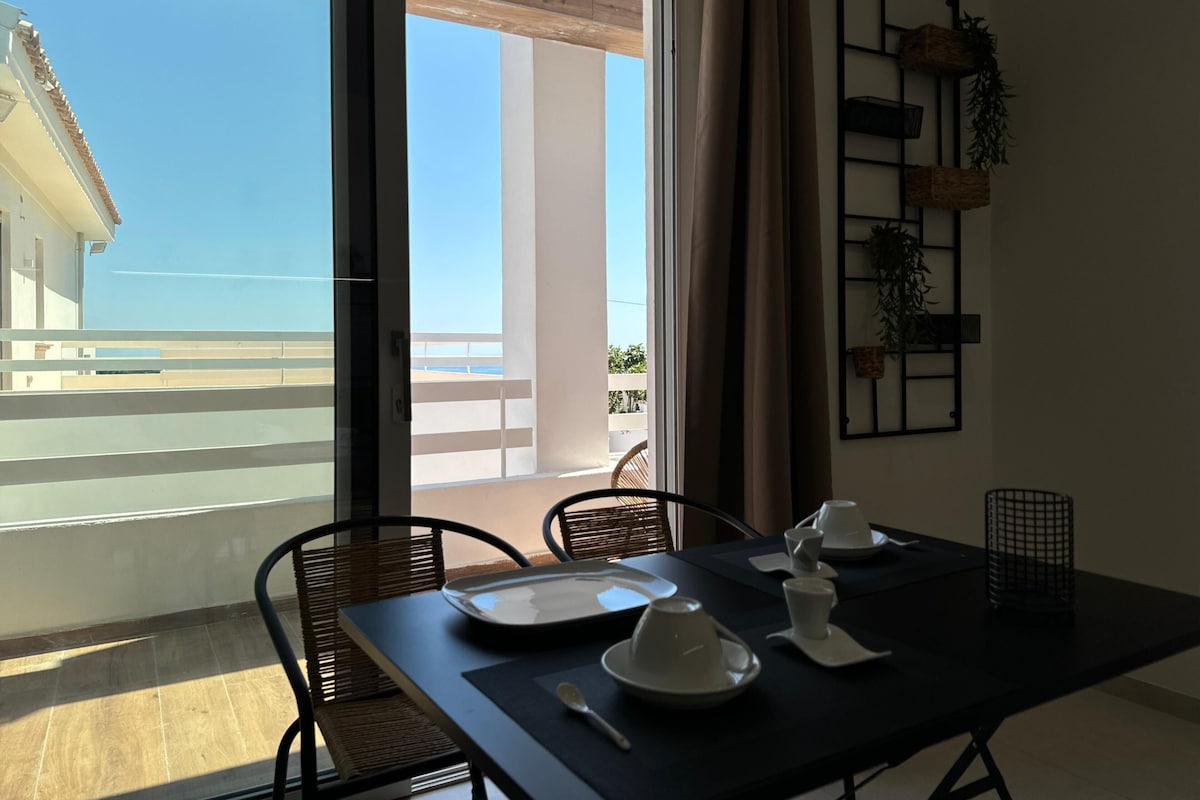 A dining area with a table set for two, a view of the balcony, and a plant on the wall.