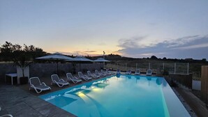 A rectangular swimming pool with lounge chairs and umbrellas, surrounded by a low stone wall, set against a sunset sky. - Itanco Resort (Crotone)
