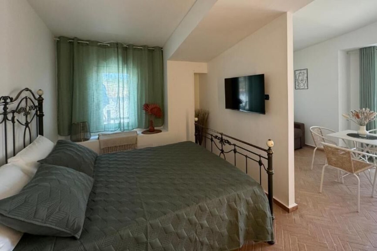 A bedroom with a bed, a TV, a balcony with a table and chairs, and a window with curtains.