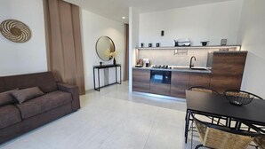 A modern kitchen with a built-in oven, a dining area with a table and chairs, a sofa, and a small side table with a vase. - Itanco Resort (Crotone)