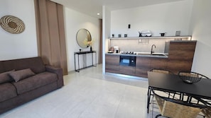 A modern kitchen with a built-in oven, a dining area with a table and chairs, a sofa, and a small side table with a vase.