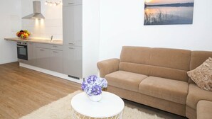 Uma cozinha moderna com forno embutido, uma área de refeições com mesa branca e um vaso de flores, e uma sala de estar com sofá bege e uma pintura de um pôr do sol.