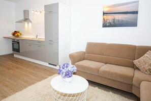 Una cucina moderna con forno integrato, una zona pranzo con tavolo bianco e un vaso di fiori, e un soggiorno con divano color beige e un quadro raffigurante un tramonto.