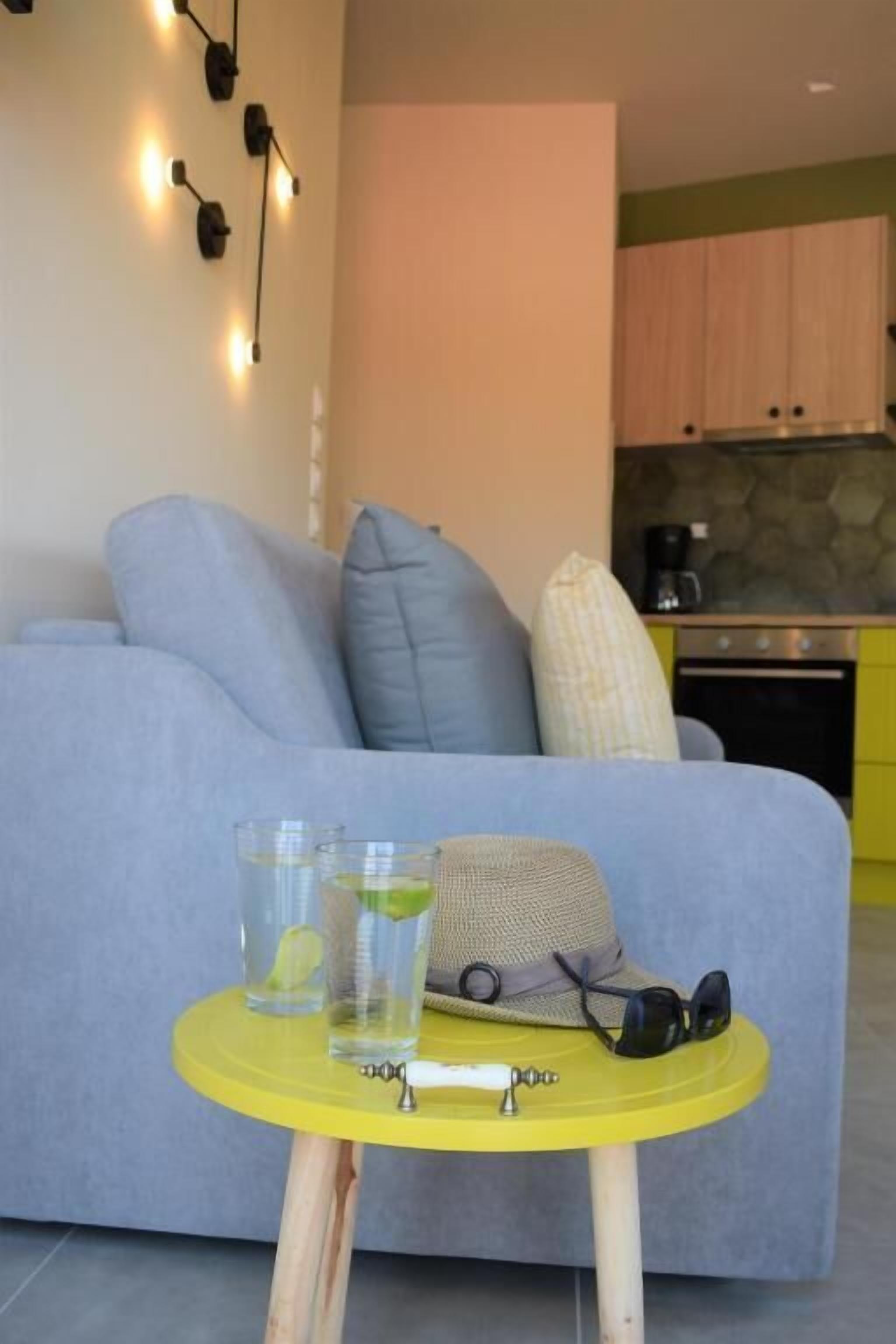 Una sala de estar con un sofá, una mesita amarilla con vasos y un sombrero, y una cocina al fondo.