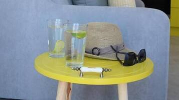Una sala de estar con un sofá, una mesita amarilla con vasos y un sombrero, y una cocina al fondo.
