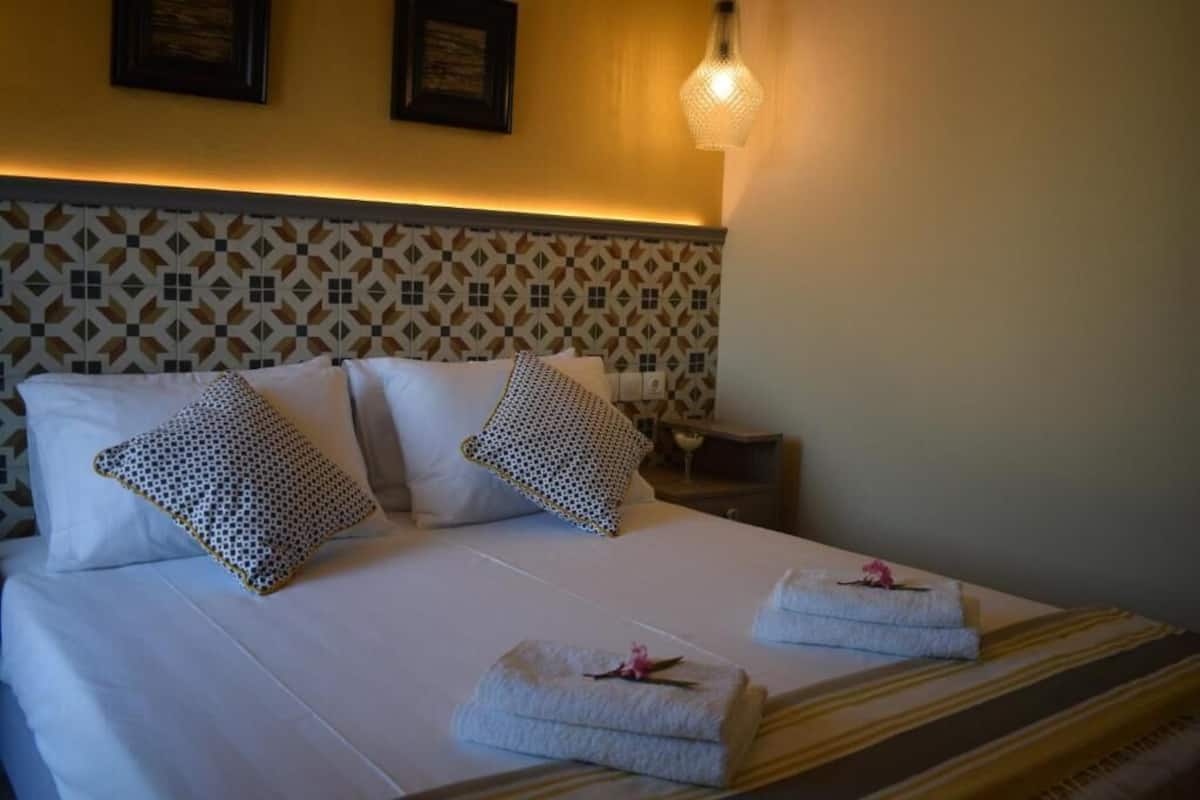 Un lit bien fait, agrémenté d’oreillers à motifs, une table de chevet avec une lampe, et des serviettes soigneusement pliées posées sur le lit.