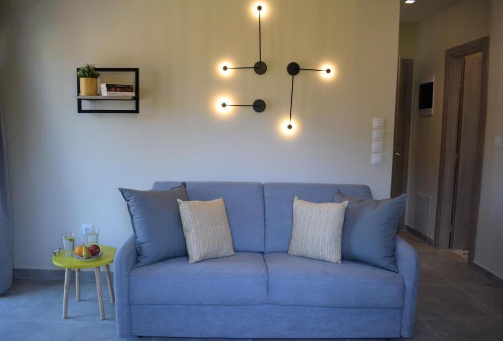 A modern living room with a grey sofa, a wall-mounted light fixture, and a small table with fruit and drinks.