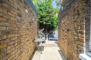 A narrow brick alleyway with a table and chairs, a hanging plant, and a tree in the background. - Stunning 2 Bedroom Apart| Private Garden | Fulham (Greater London)