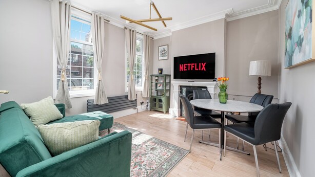Ein Wohnzimmer mit einem grünen Sofa, einem Esstisch mit Stühlen, einem Kamin und einem Fernseher, der 'NETFLIX' anzeigt.