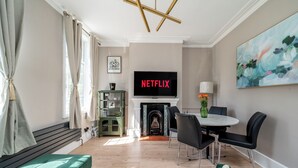Ein Wohnzimmer mit Kamin, einem Fernseher, der 'NETFLIX' zeigt, einer Essecke mit Stühlen und einem grünen Sofa.