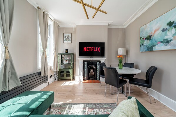A living room with a fireplace, a television displaying 'NETFLIX,' a dining area with chairs, and a green sofa. - Stunning 2 Bedroom Apart| Private Garden | Fulham (Greater London)