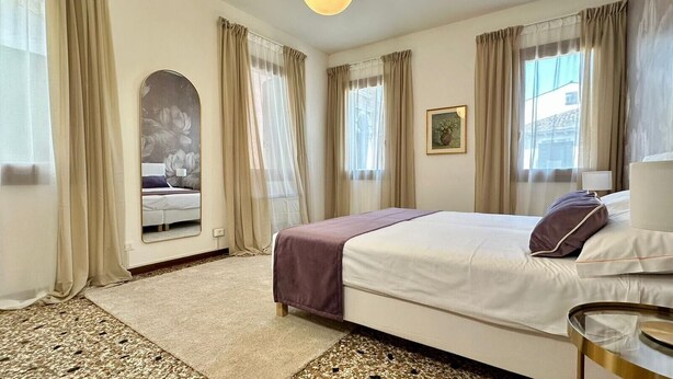 Ein Hotelzimmer mit Bett, Spiegel, einem fensterbepflanzten Fenster, Teppich und kleinem Tisch mit Lampe.
