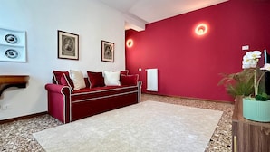 Ein rotes Sofa mit weißen und roten Kissen, ein weißer Teppich, ein Holznachttisch mit einer Pflanze und gerahmte Bilder an der Wand.