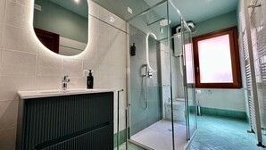 Ein modernes Badezimmer mit einer gläsernen Duschkabine, einem weißen Waschbecken inklusive Seifenspender und einer durch die Glastür erkennbaren Toilette.