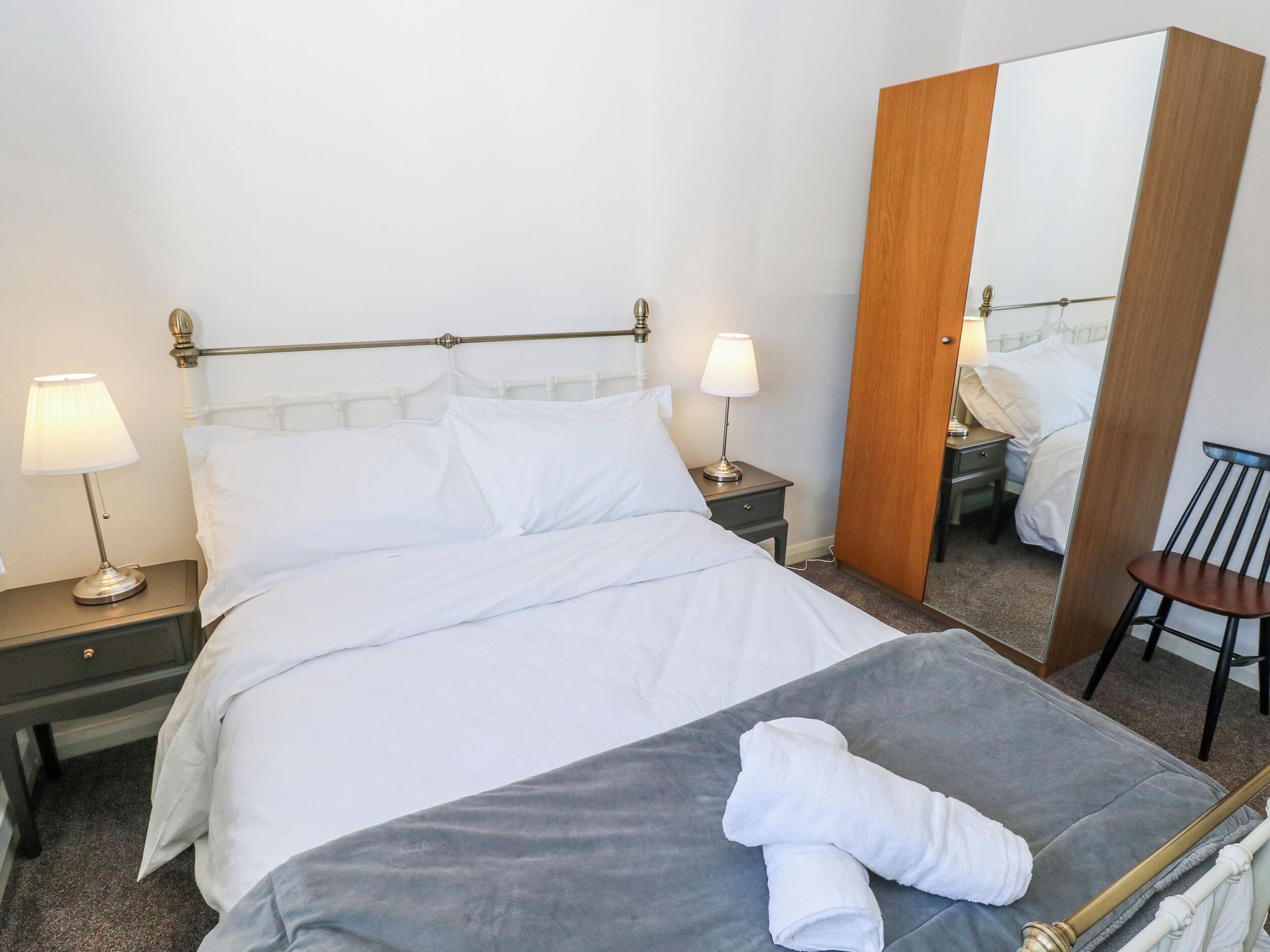 A neatly made bed with white linens, two bedside lamps, a wooden door, and a chair.