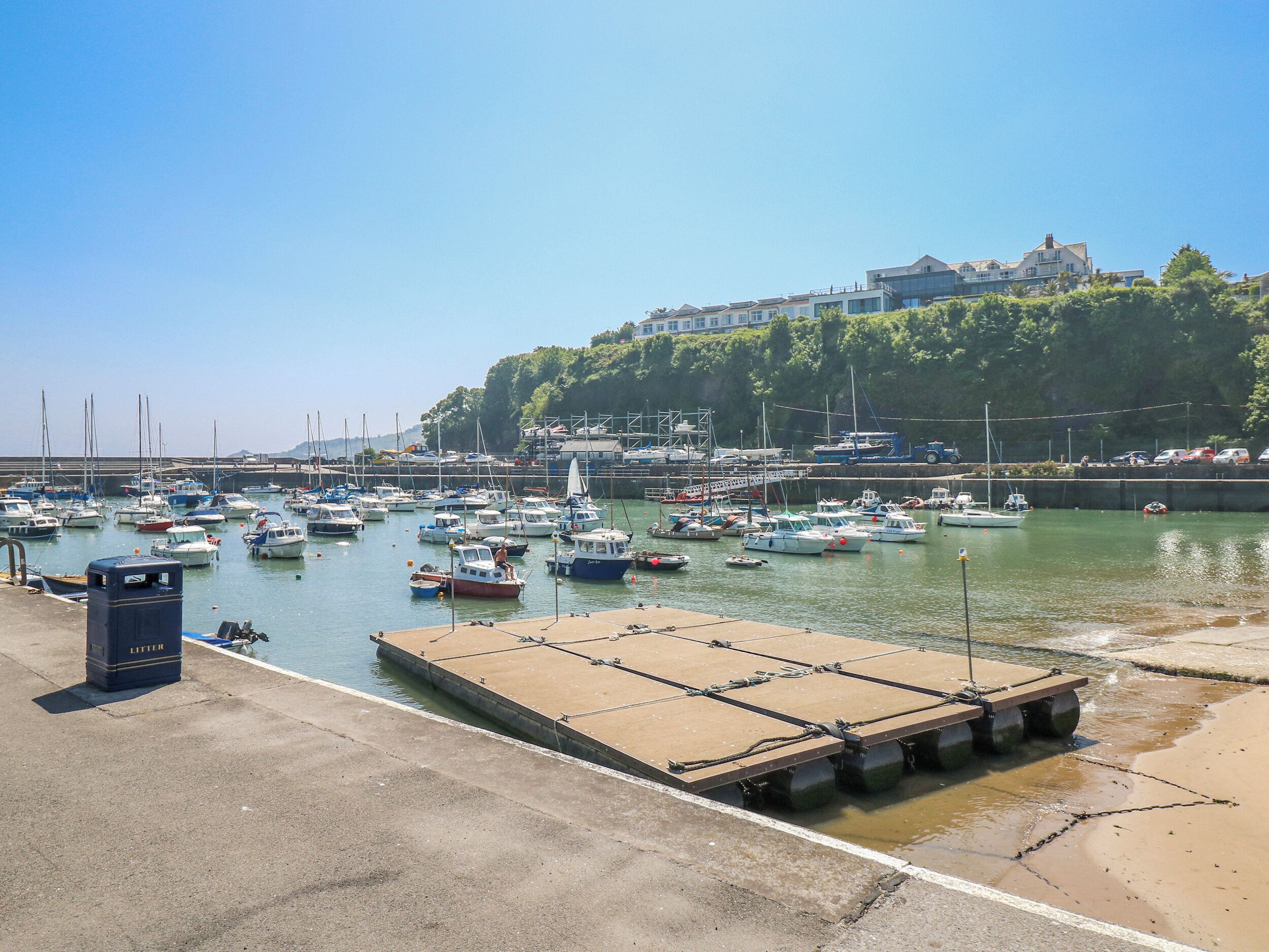 A marina with boats docked, a concrete pier, and a building with a hill in the background.