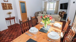 A dining area with a wooden table set for four, chairs, a vase with flowers, and a television in the background. - Moonraker (Saundersfoot)