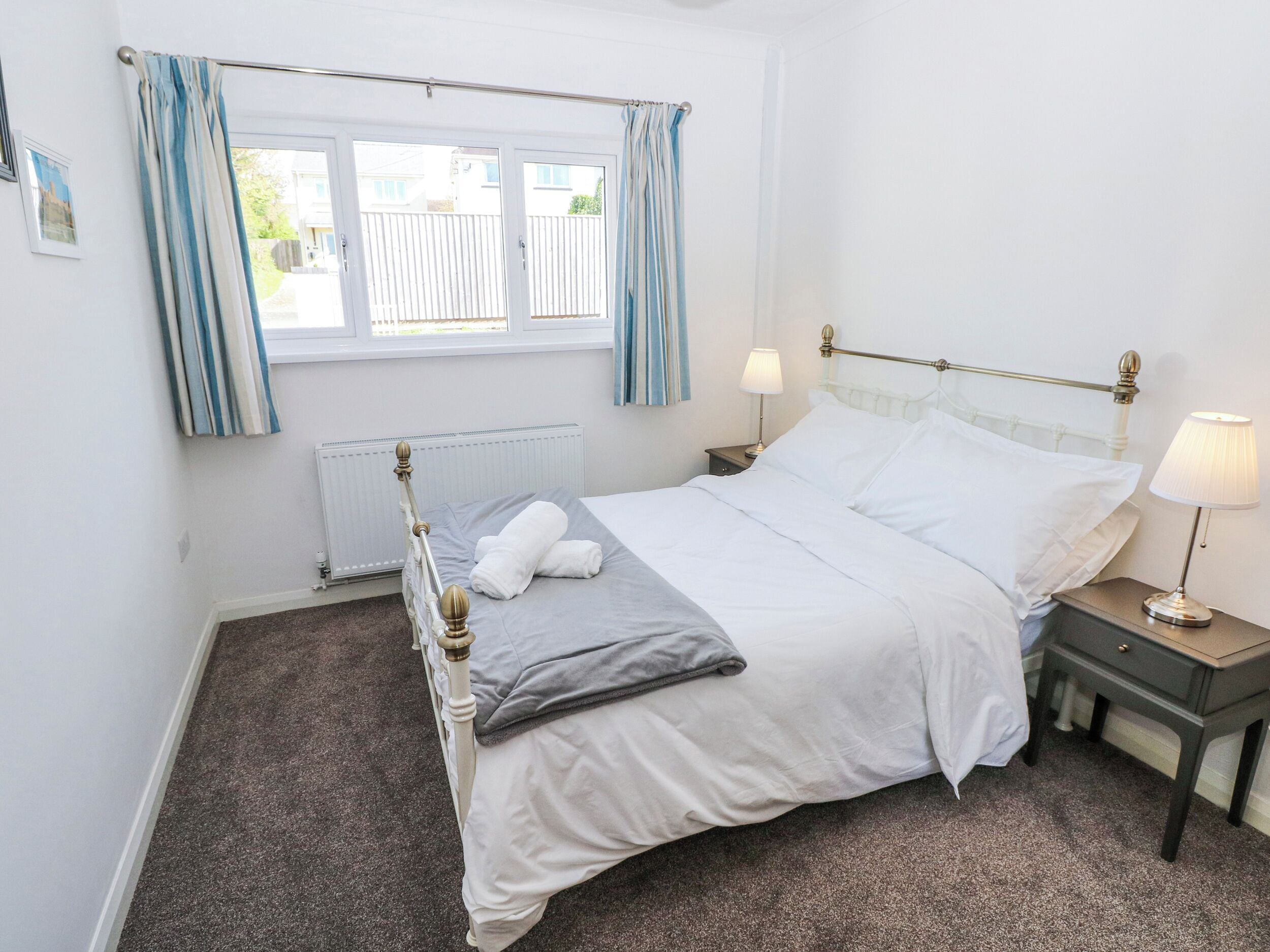 A bedroom with a bed, two bedside tables, a window with curtains, a radiator, and a wall-mounted picture.