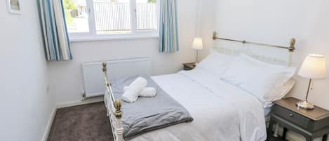 A bedroom with a bed, two bedside tables, a window with curtains, a radiator, and a wall-mounted picture.