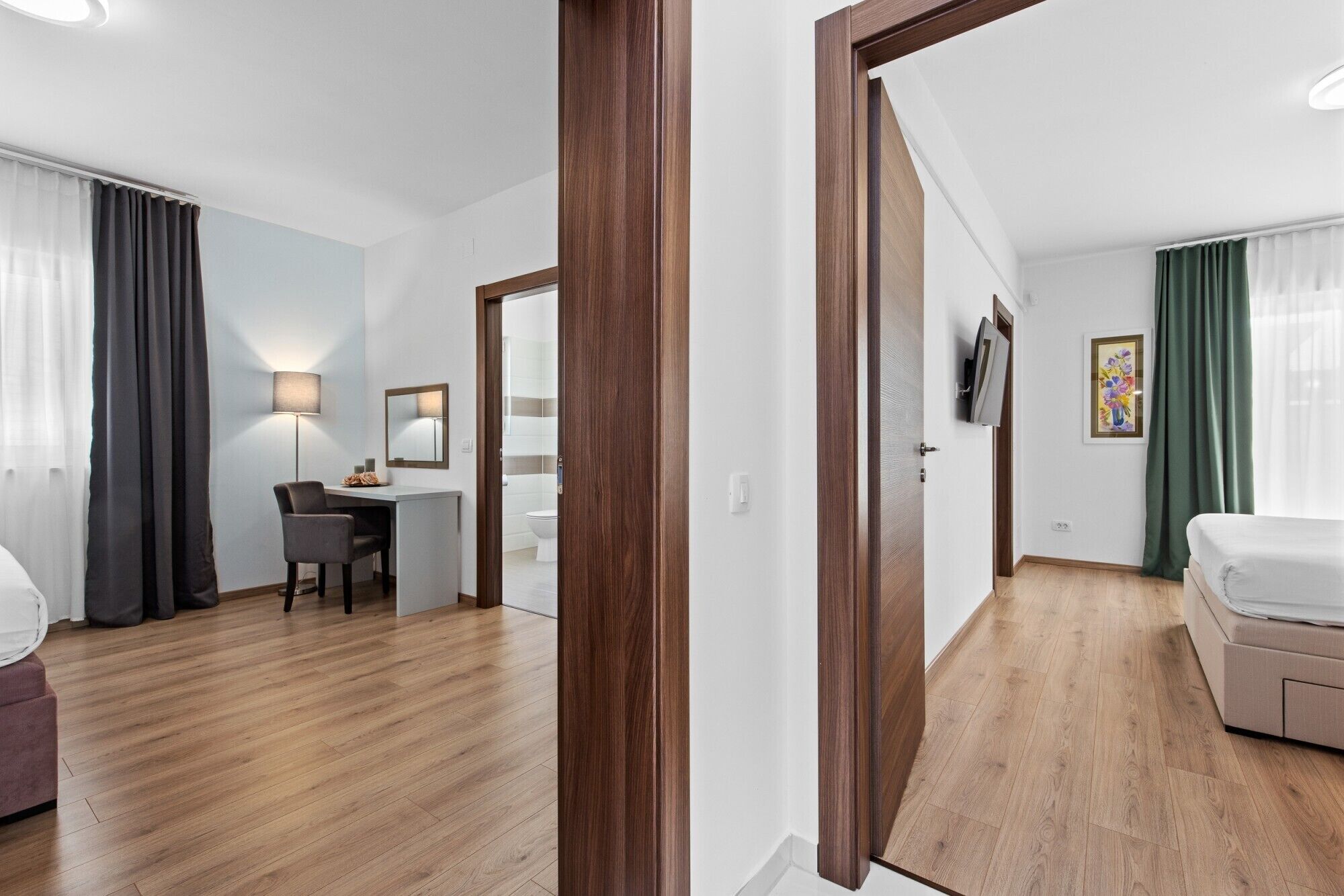 Habitación de hotel con piso de madera, un escritorio con silla, una cama, un televisor y un baño visible a través de una puerta abierta.