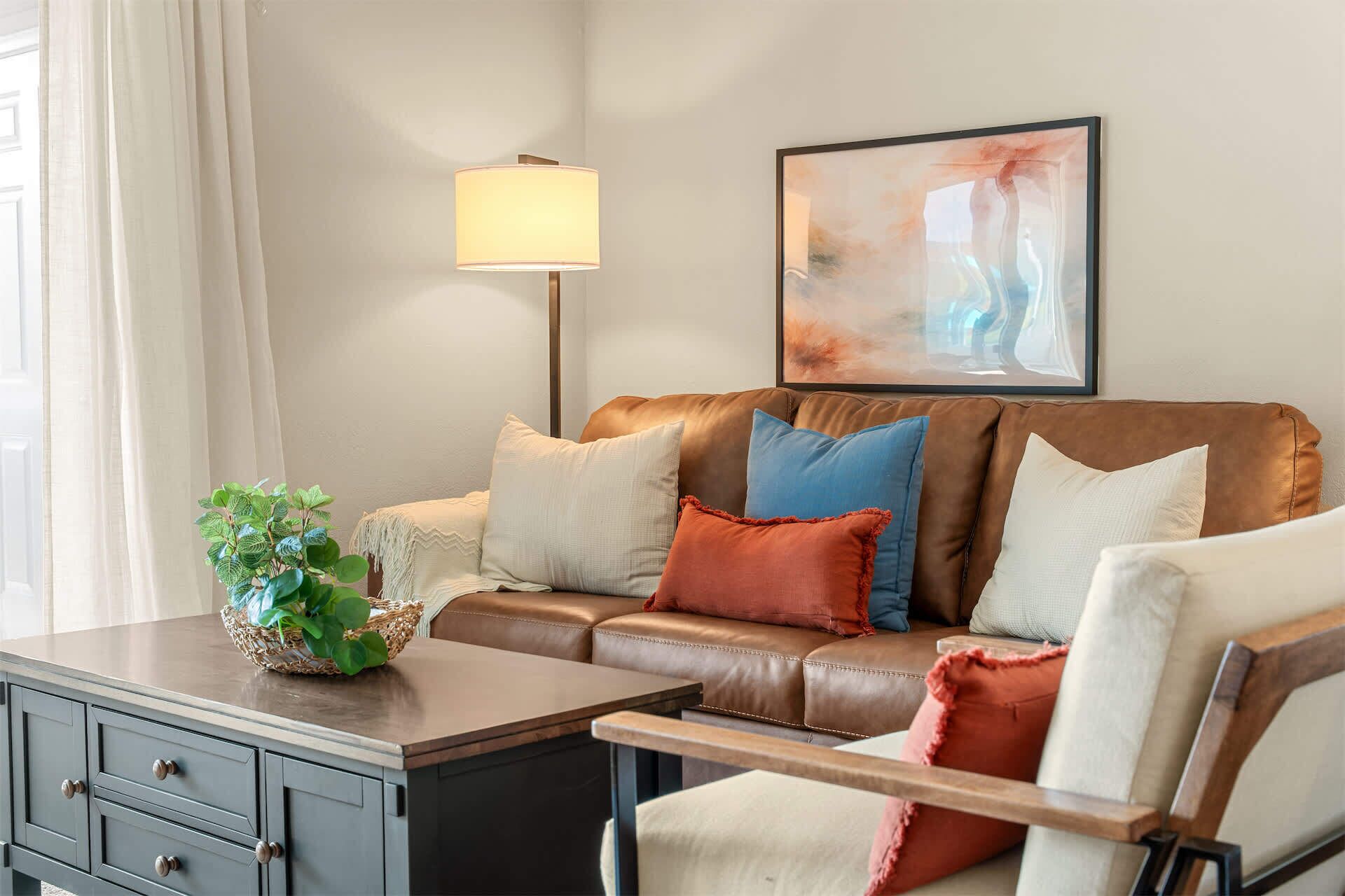 A living room with a brown leather sofa, a side table with a plant, a lamp, and a framed abstract painting on the wall.