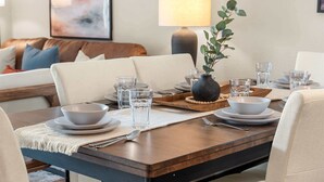 Uma área de jantar com uma mesa posta para quatro pessoas, um vaso com folhagem e um abajur ao fundo.