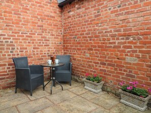 A corner patio with two wicker chairs, a small round table, and two potted plants. - 1 The Stables (Wrexham)