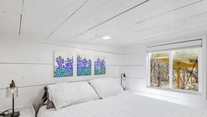 A bedroom with a bed, white walls, a window with a view, and three paintings on the wall. - Charming Tiny Home Retreat | Porch Swing, Grill, Stargazing Near Caverns (Bulverde)