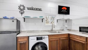 A kitchen with a washing machine, a refrigerator, a sink, and a TV mounted on the wall displaying 'HCPL Hill Country Premier Leading.' - Charming Tiny Home Retreat | Porch Swing, Grill, Stargazing Near Caverns (Bulverde)