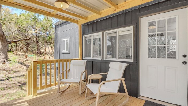 A wooden deck with white rocking chairs and a white door with a 'welcome' sign. - Charming Tiny Home Retreat | Porch Swing, Grill, Stargazing Near Caverns (Bulverde)