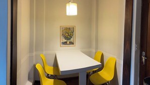 A dining area with a white table and four yellow chairs, a hanging light fixture, and a framed painting on the wall. - Apartment in the Tourist Center with 24-hour Reception/Concierge (Campos do Jordão)