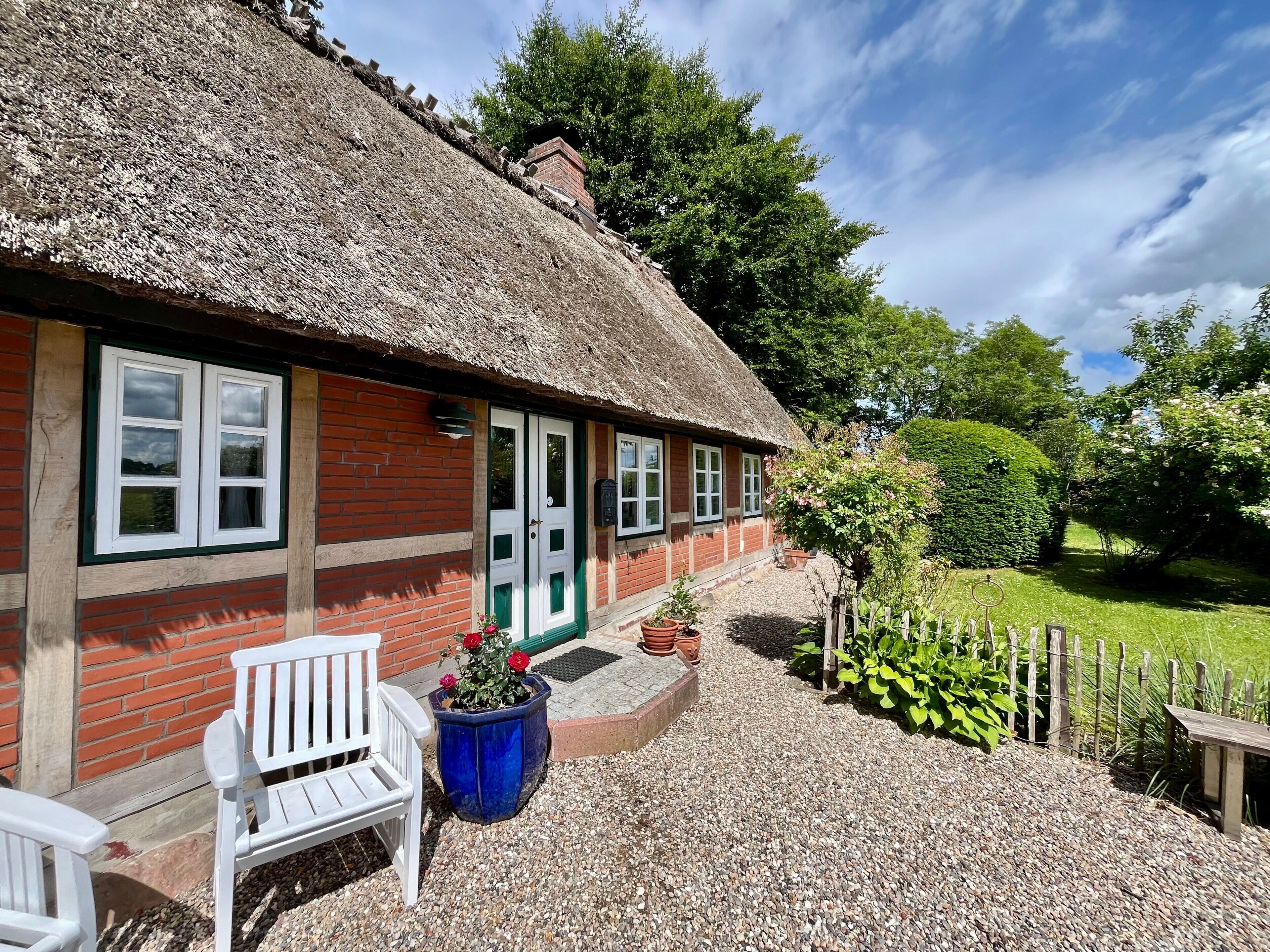 Ein reetgedecktes Cottage mit Ziegelaußenwand, Fenstern mit weißen Rahmen und einer Schotterterrasse mit einem weißen Stuhl und Pflanzkübeln.