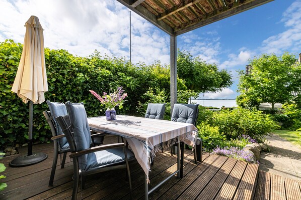 An outdoor patio with a table and chairs, surrounded by greenery and a view of the sea.