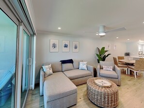A modern living room with a sectional sofa, a round woven ottoman, and a dining area with a table and chairs. - Pointe South 104 | Gulf-Front, Screened Lanai, Pool Access + Steps to the Beach (Fort Myers Beach)
