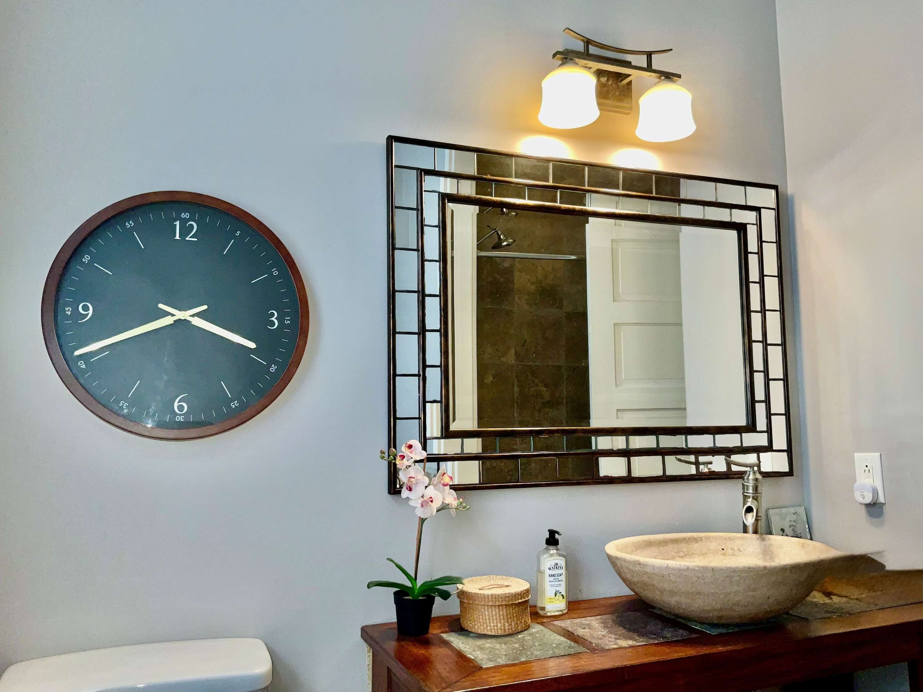 A bathroom with a round wall clock, a mirror, a sink, and a small potted orchid.