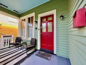 A porch with a red door, two chairs, a small table, and a striped rug. - Verdant Red Door Retreat (Galveston)