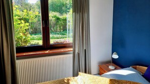 Ein Schlafzimmer mit großem Fenster, Heizkörper, Nachttisch mit Lampe und Bett mit gesteppter Decke.
